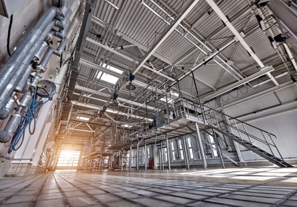 Perspektywiczne ujęcie wnętrza nowoczesnej hali przemysłowej o wysokim stropie. Widoczne są liczne instalacje rur, metalowe podesty ze schodami oraz systemy oświetleniowe, a przez otwarte wrota na końcu hali wpada jasne, naturalne światło.
