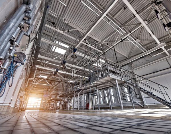 Perspektywiczne ujęcie wnętrza nowoczesnej hali przemysłowej o wysokim stropie. Widoczne są liczne instalacje rur, metalowe podesty ze schodami oraz systemy oświetleniowe, a przez otwarte wrota na końcu hali wpada jasne, naturalne światło.